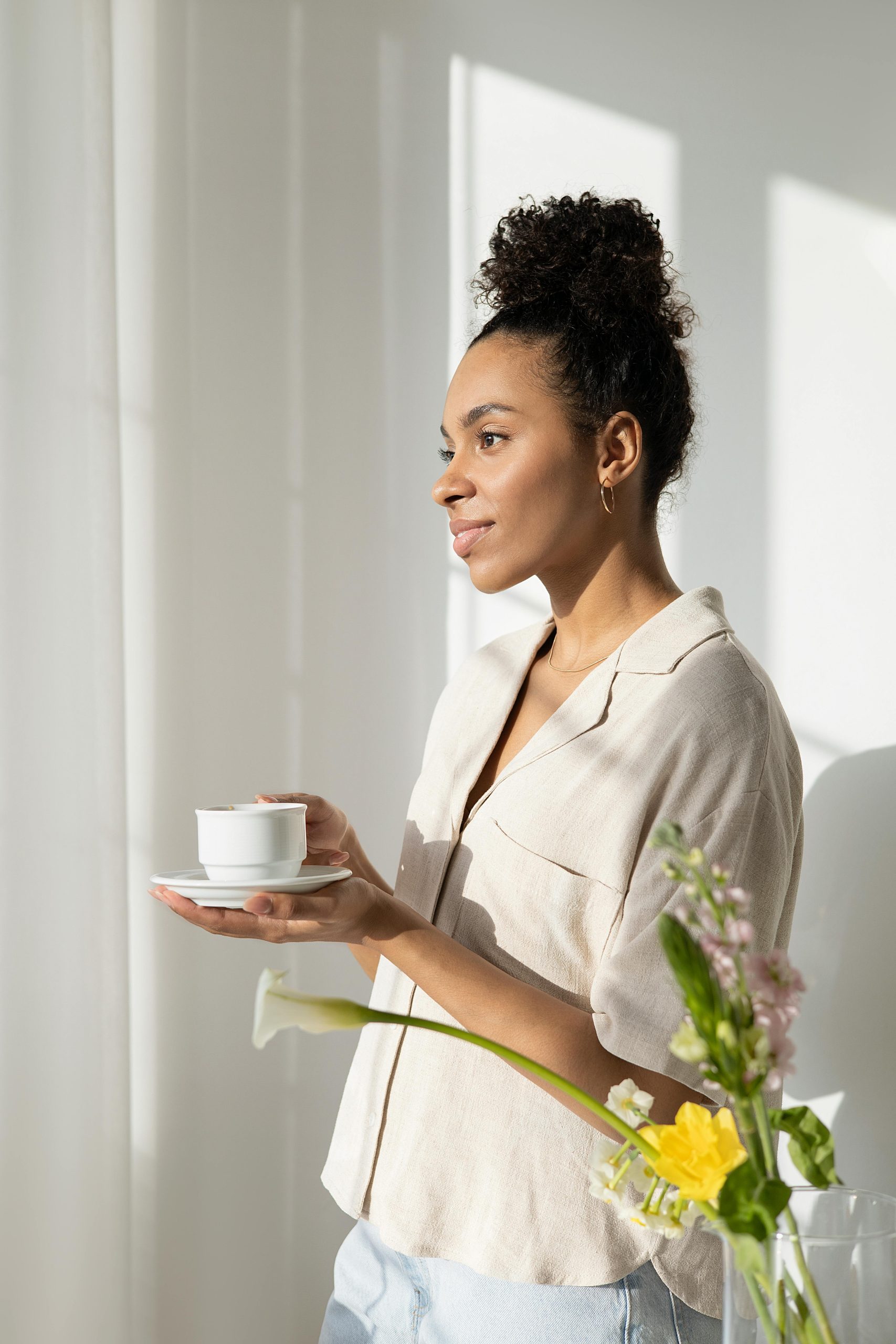 A lady stands in the window with a cup of tea in her hand and flowers next to her