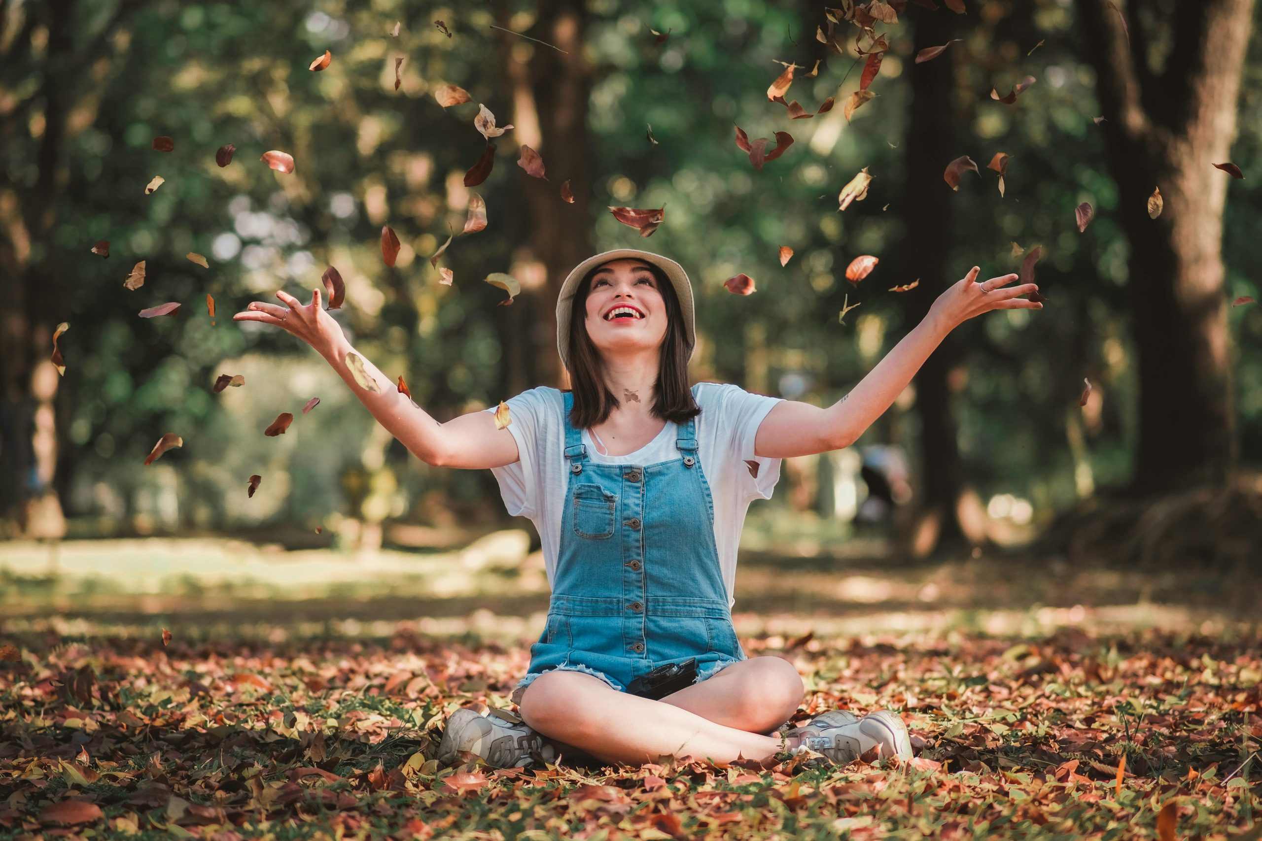 Young lady sits under trees throwing autumn leaves in the air