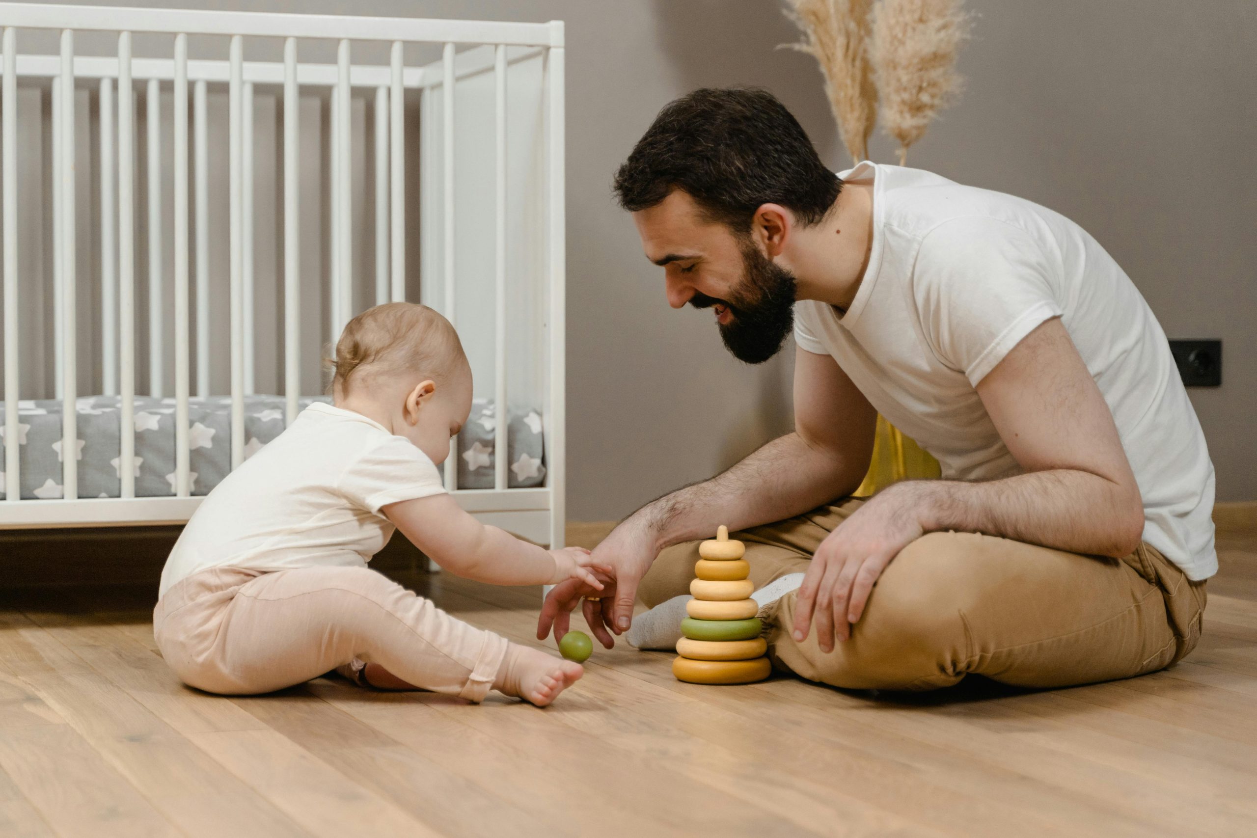 Man playing game with a baby sitting crossed legged on the floor in front of cot