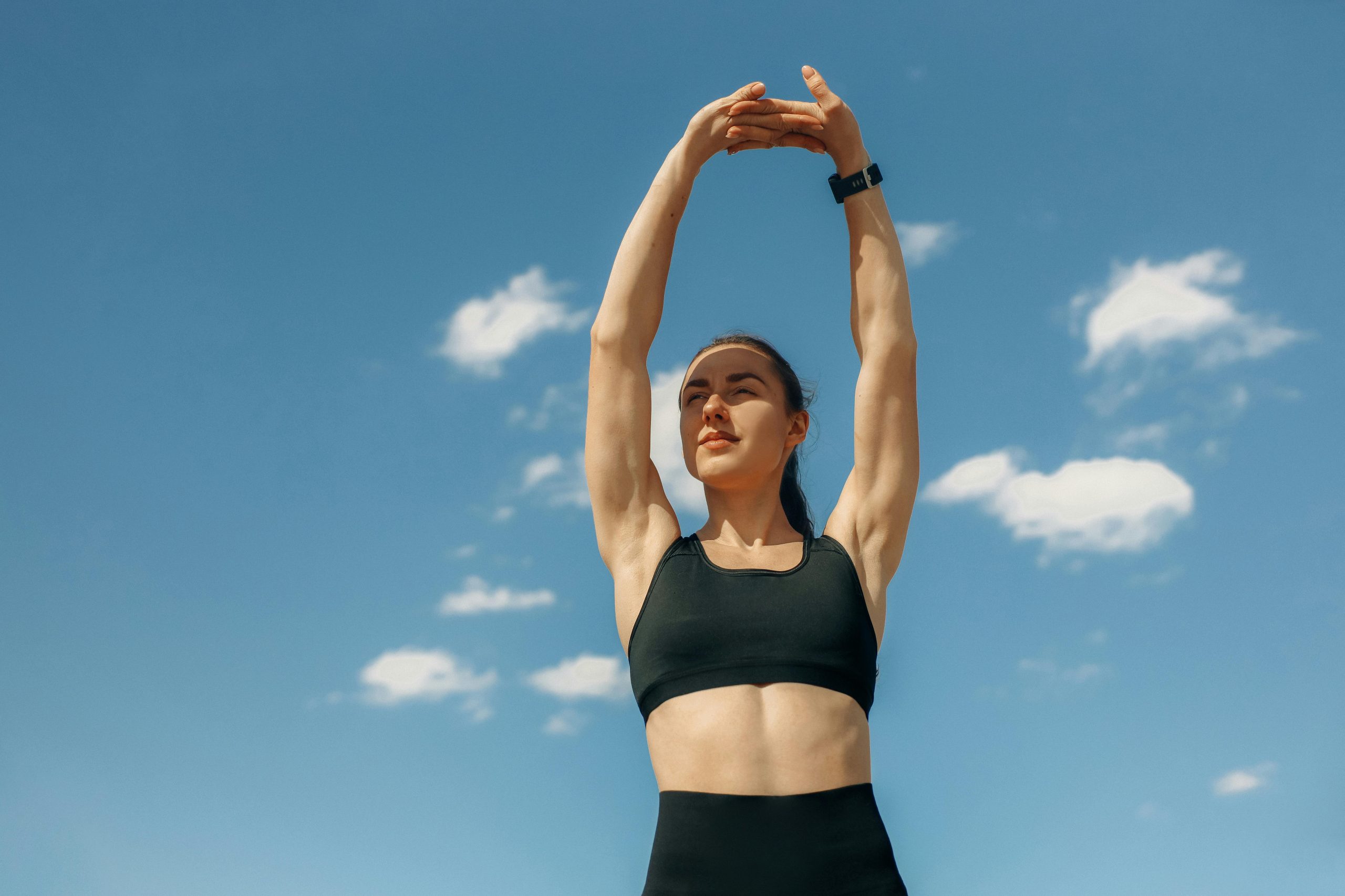 Sports woman in crop top stretching shoulders with blue sky and clouds behind